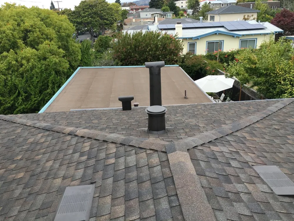 A rooftop view showcasing a shingle roof with vent pipes and chimneys, surrounded by trees and residential homes in Oakland.